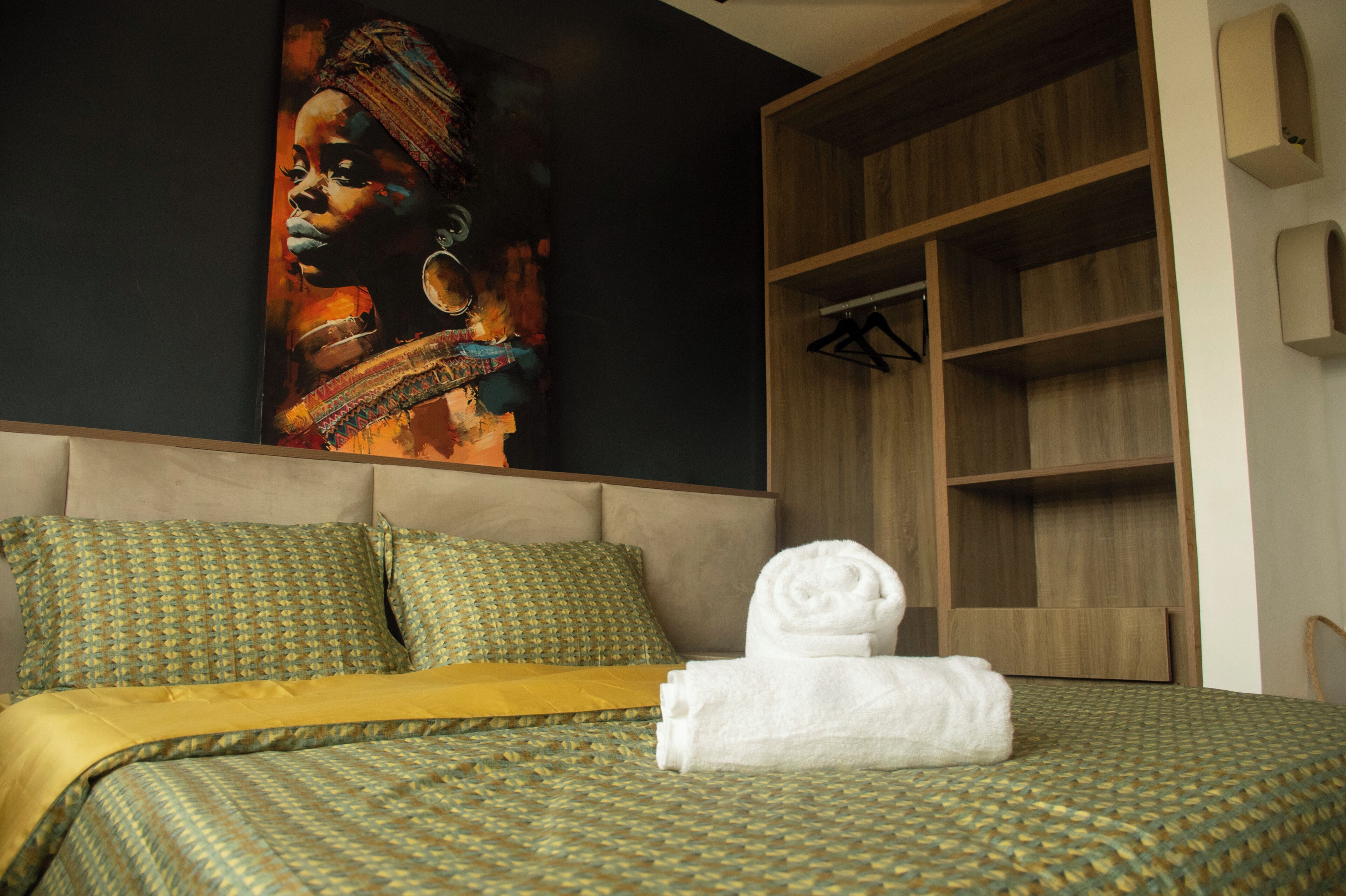 Quarto de hotel com uma cama, um quadro grande na parede, um guarda-roupa de madeira e uma cama bem arrumada com uma toalha branca enrolada.