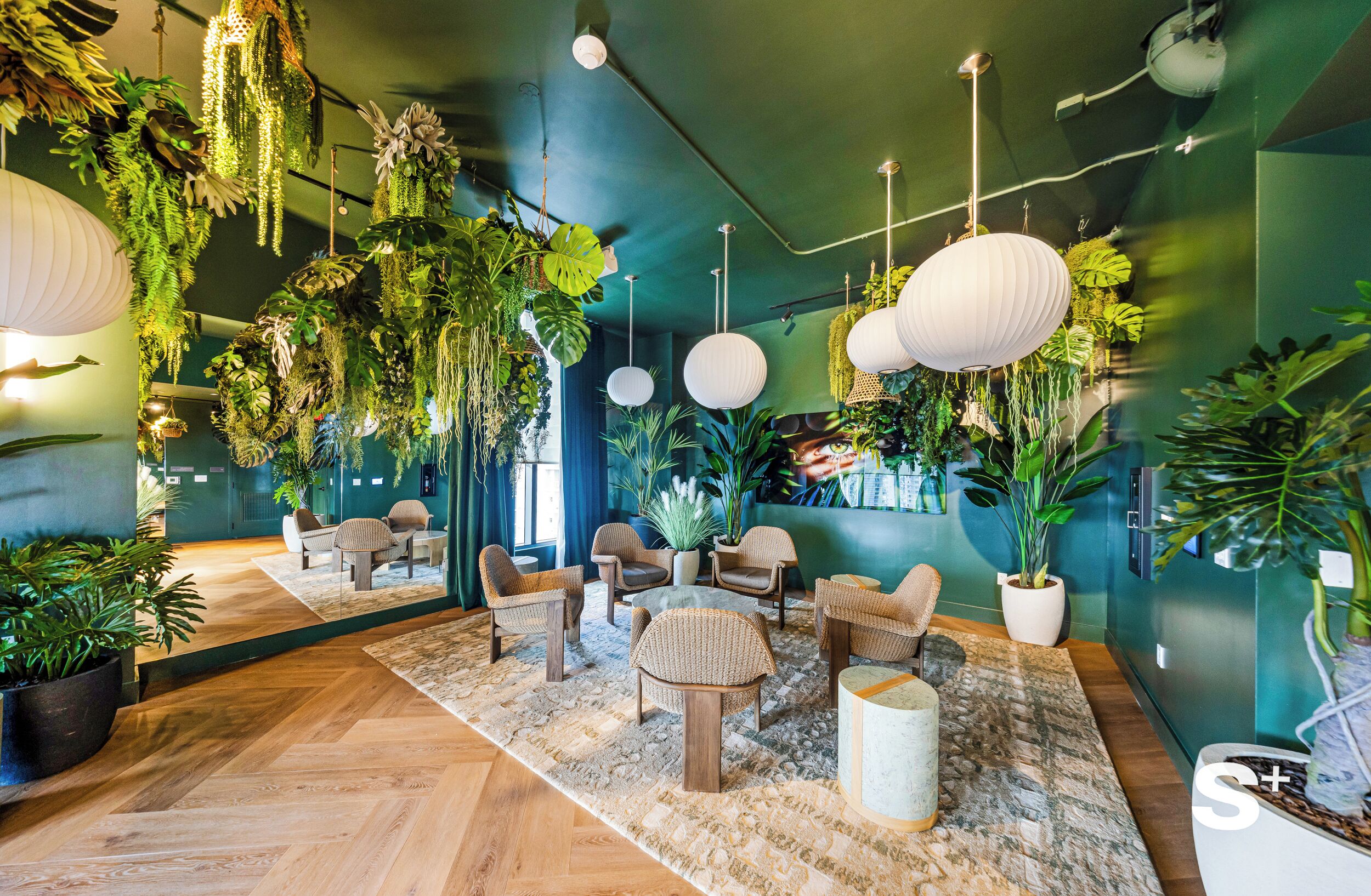 A room with a herringbone floor, wicker furniture, hanging plants, and white paper lanterns.
