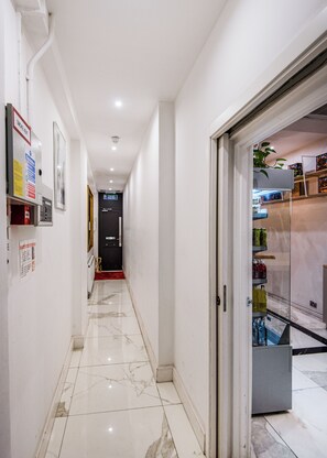 A hallway with a marble floor, a fire alarm, and a door leading to a room with a refrigerator and shelves. - SomersetHouseSuites by Aureva Residences (London)