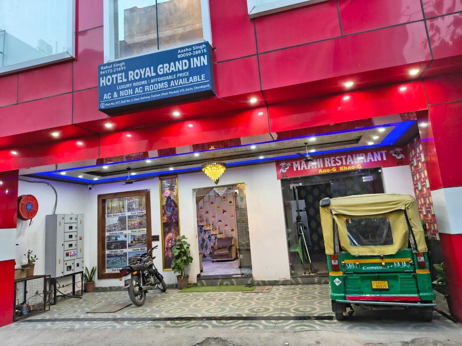 La entrada de un hotel con un letrero que dice Hotel Royal Grand Inn, una motocicleta estacionada afuera y un auto-rickshaw verde adelante.
