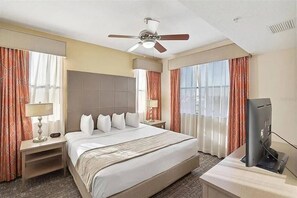Ein Hotelzimmer mit einem Bett, einem Schreibtisch mit Fernseher, einem Deckenventilator und großen Fenstern mit Vorhängen.