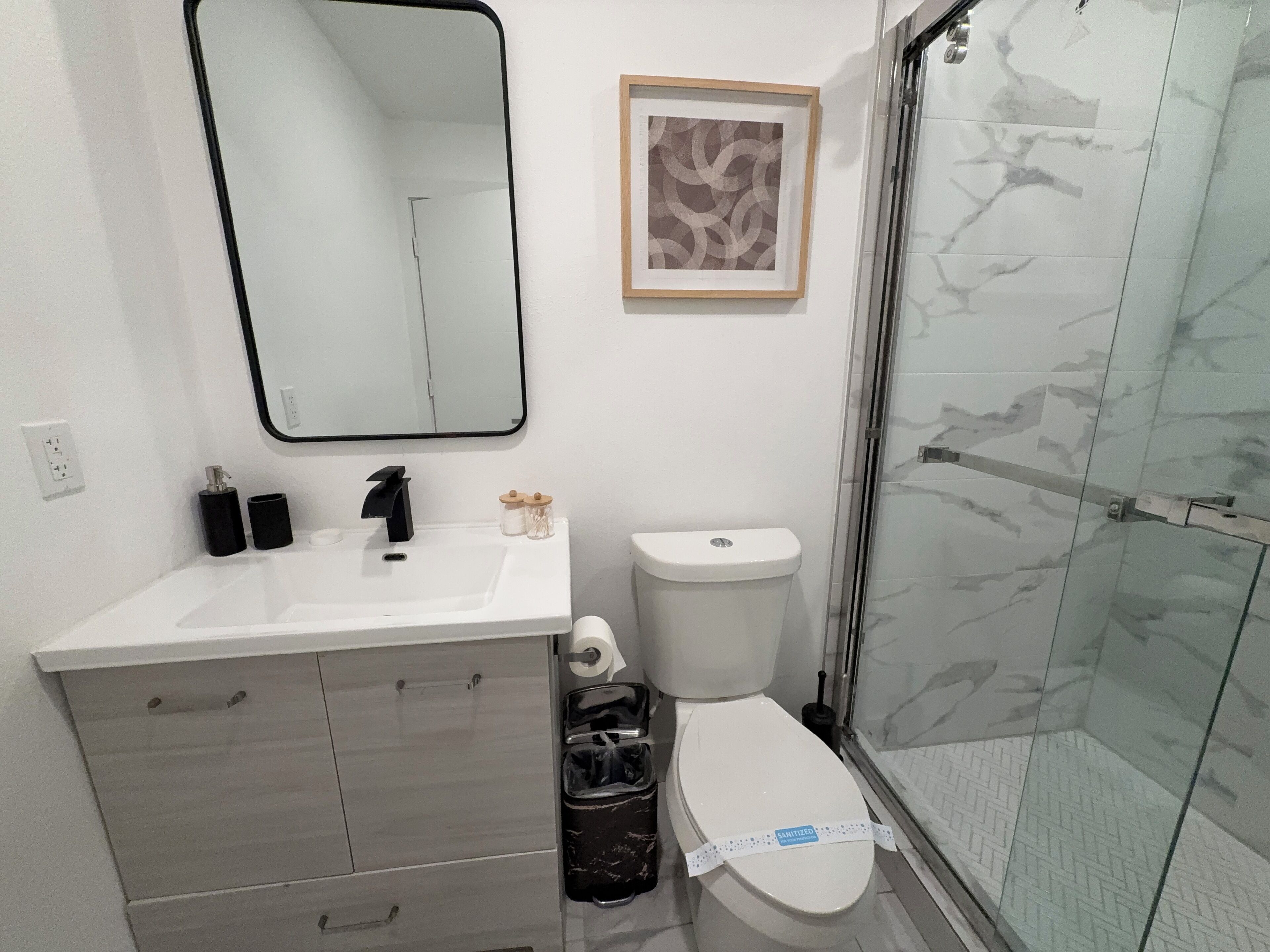 A bathroom with a white sink, black faucet, a framed picture, a toilet, and a glass shower enclosure.