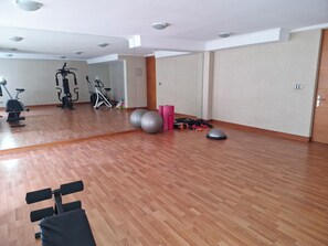 Ein geräumiges Zimmer mit Holzboden, ausgestattet mit Fitnessgeräten wie einem Ergometer, Crosstrainer und Gewichten.