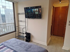 A hotel room with a bed, a TV mounted on the wall, a window with a view of buildings, and a wooden door. - Renovated Apartment in the Heart of Providencia Balcony & A/C (Providencia)