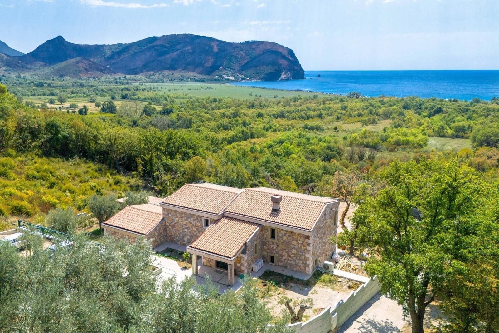 Une maison en pierre avec un toit de tuiles, entourée d’une végétation luxuriante et offrant une vue sur la mer et les montagnes.