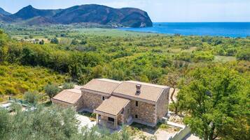 Une maison en pierre avec un toit de tuiles, entourée d’une végétation luxuriante et offrant une vue sur la mer et les montagnes.