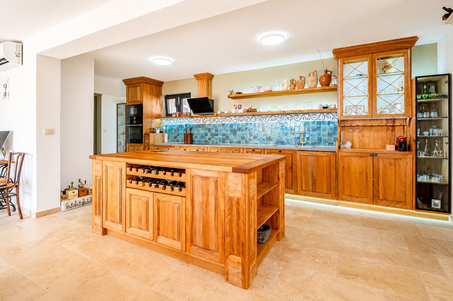 Une cuisine avec des meubles en bois, un îlot central doté d’un espace de rangement pour les bouteilles de vin, un dosseret carrelé et un four encastré.
