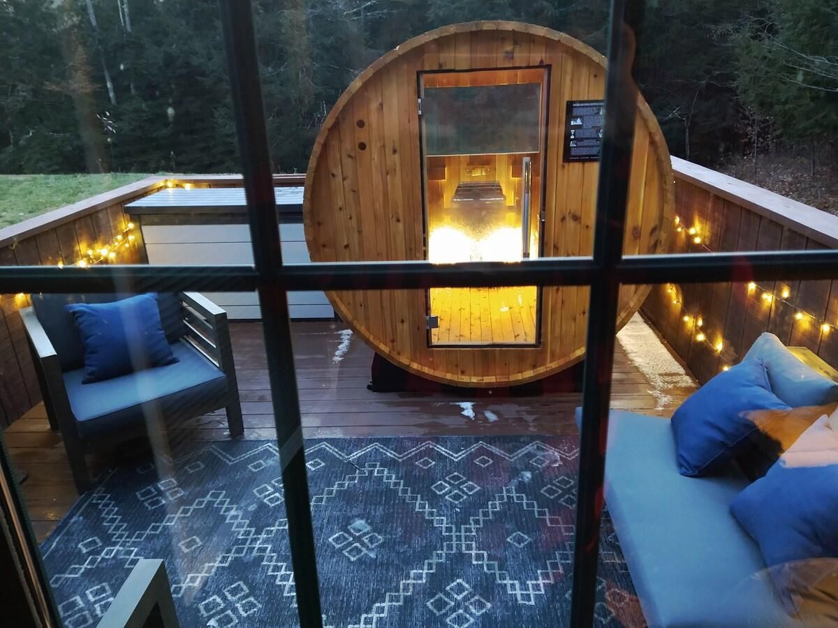 Uma cabana de madeira com estrutura de vidro, um poço de fogo e um deck com assentos.