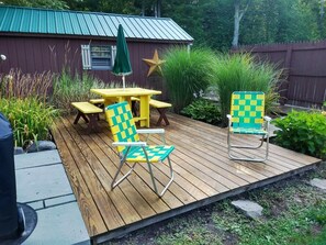 Eine Holzterrasse mit Picknicktisch und zwei karierten Stühlen, umgeben von Grünpflanzen und einem Metallzaun.
