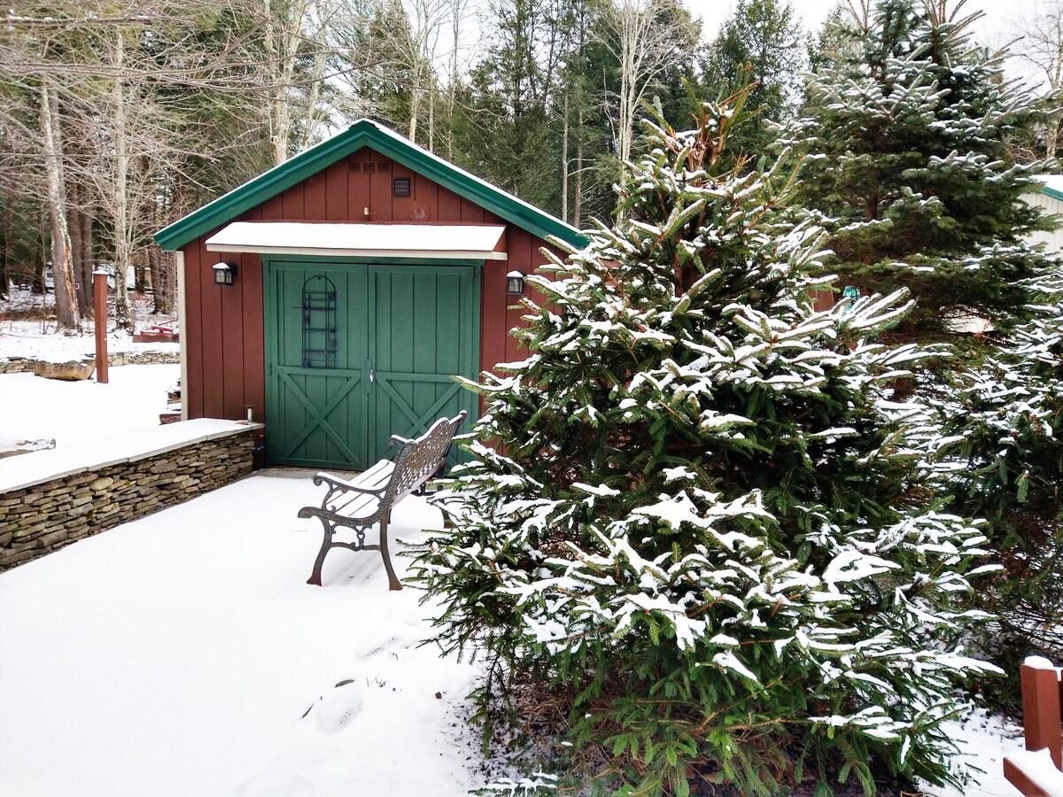 Un espacio exterior cubierto de nieve con un cobertizo de madera, un banco y un árbol de hoja perenne cargado de nieve.
