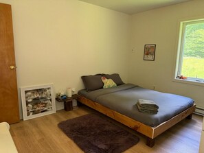 Ein Schlafzimmer mit einem Einzelbett, einem Holz-Kopfteil, einem kleinen Nachttisch, einem gerahmten Bild und einem Fenster mit Blick auf Grünflächen.