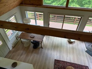 Ein Zimmer mit einem Holztisch, einem weißen Stuhl und einem Teppich. Fenster bieten Blick auf eine Holzterrasse und viel Grün.