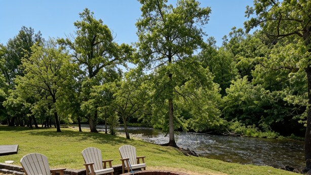 Un foyer circulaire entouré de chaises, installé sur une terrasse en béton, avec une rivière et des arbres en arrière-plan.