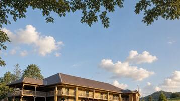 A large, two-story building with a flat roof and multiple balconies, surrounded by a grassy area and trees.