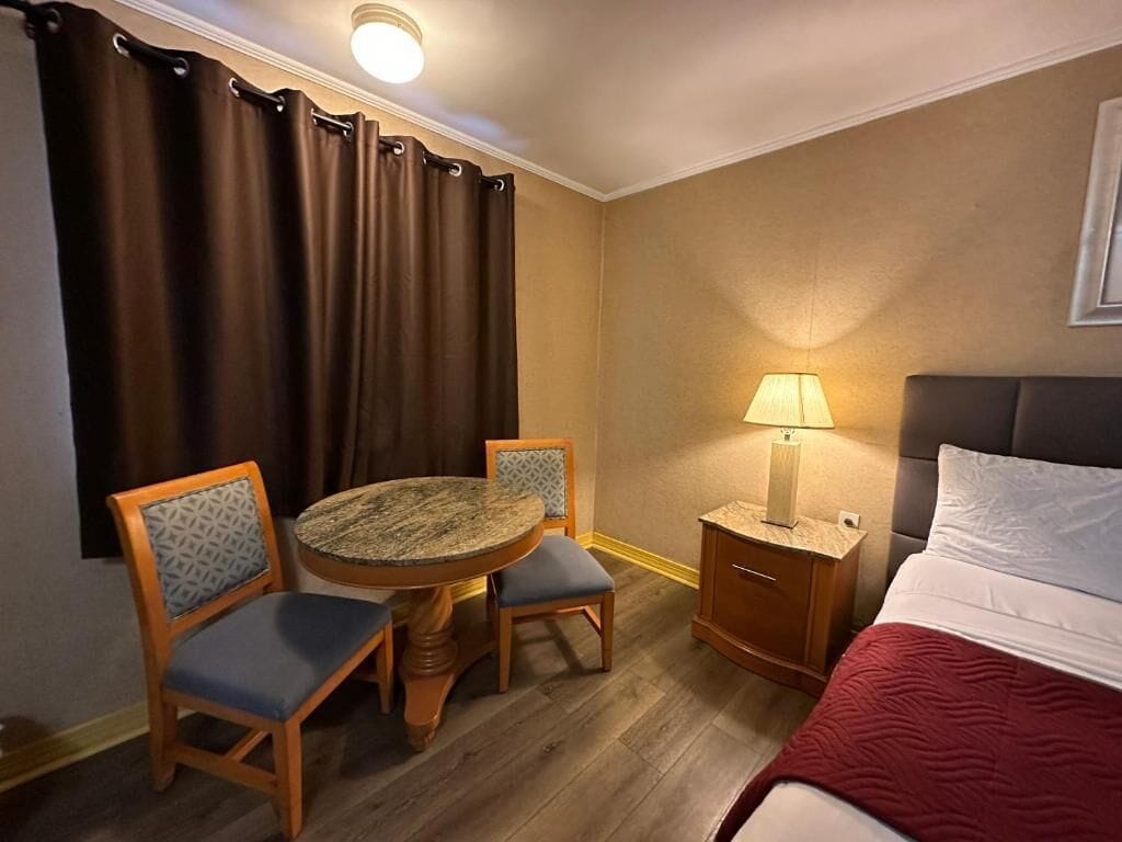 Hotellihuone, jossa on sänky, pyöreä pöytä tuoleineen, yöpöytä lampun kanssa ja ikkuna, jossa on verhot.