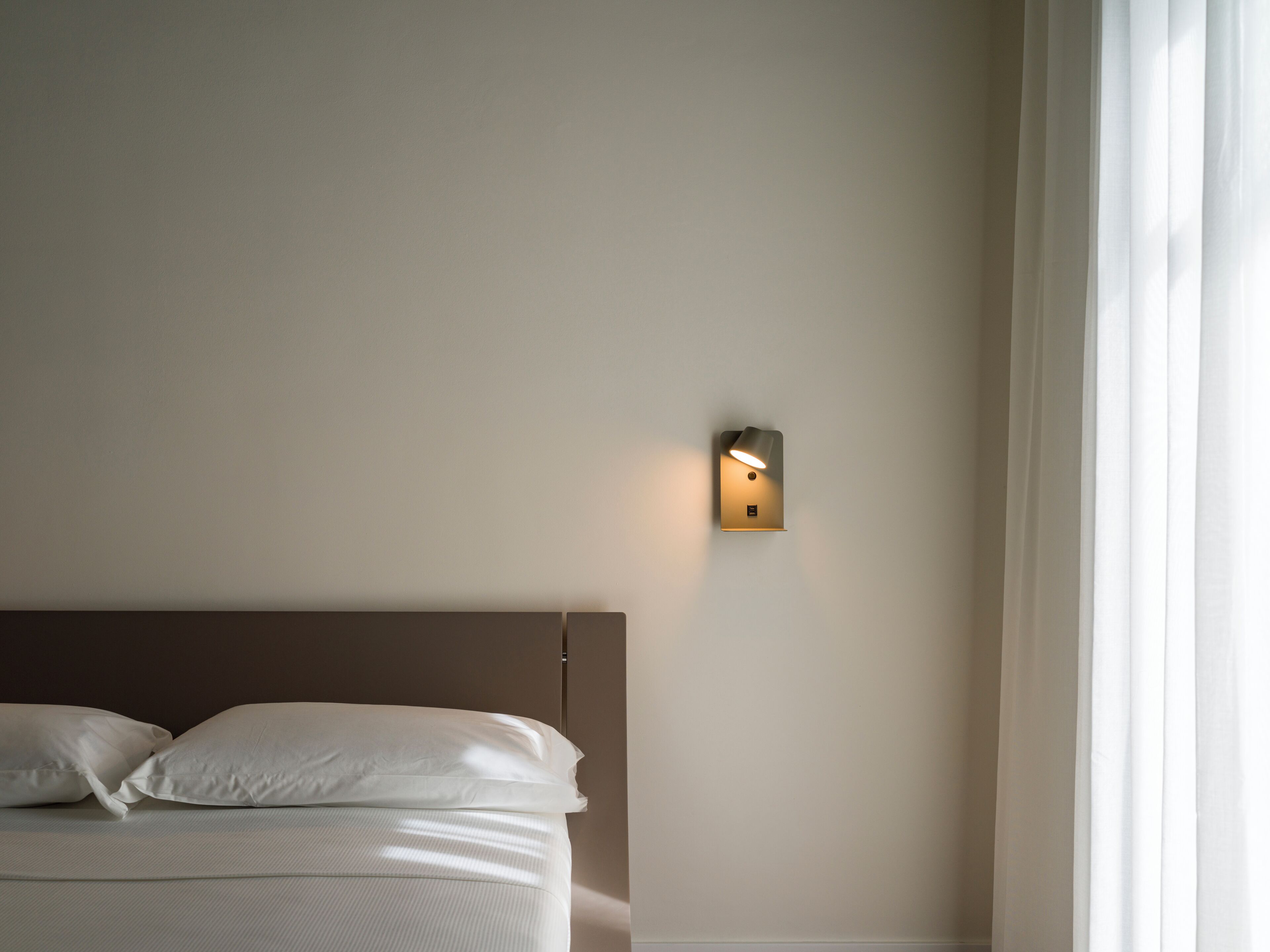 Una camera da letto con un letto, cuscini, una lampada a parete e una finestra con tende.