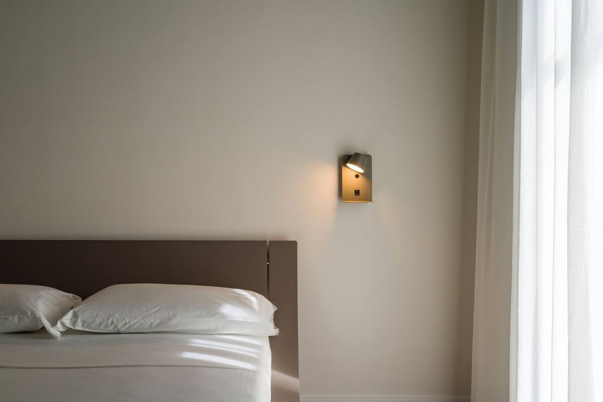 Un dormitorio con cama, almohadas, lámpara fijada a la pared y ventana con cortinas.