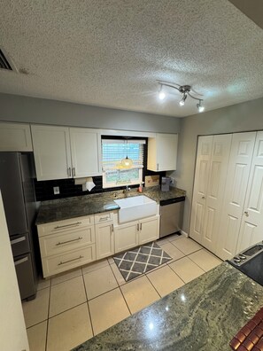 A kitchen with white cabinets, a granite countertop, a sink, a refrigerator, and a window with blinds. - Updated 3-bedroom POOL house with AC in gorgeous Fort Myers (Fort Myers)
