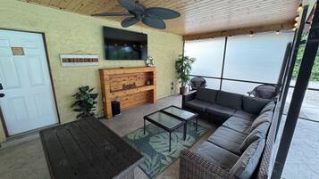 Un porche cubierto con ventilador de techo, un banco de madera, un sofá y un televisor montado en la pared.