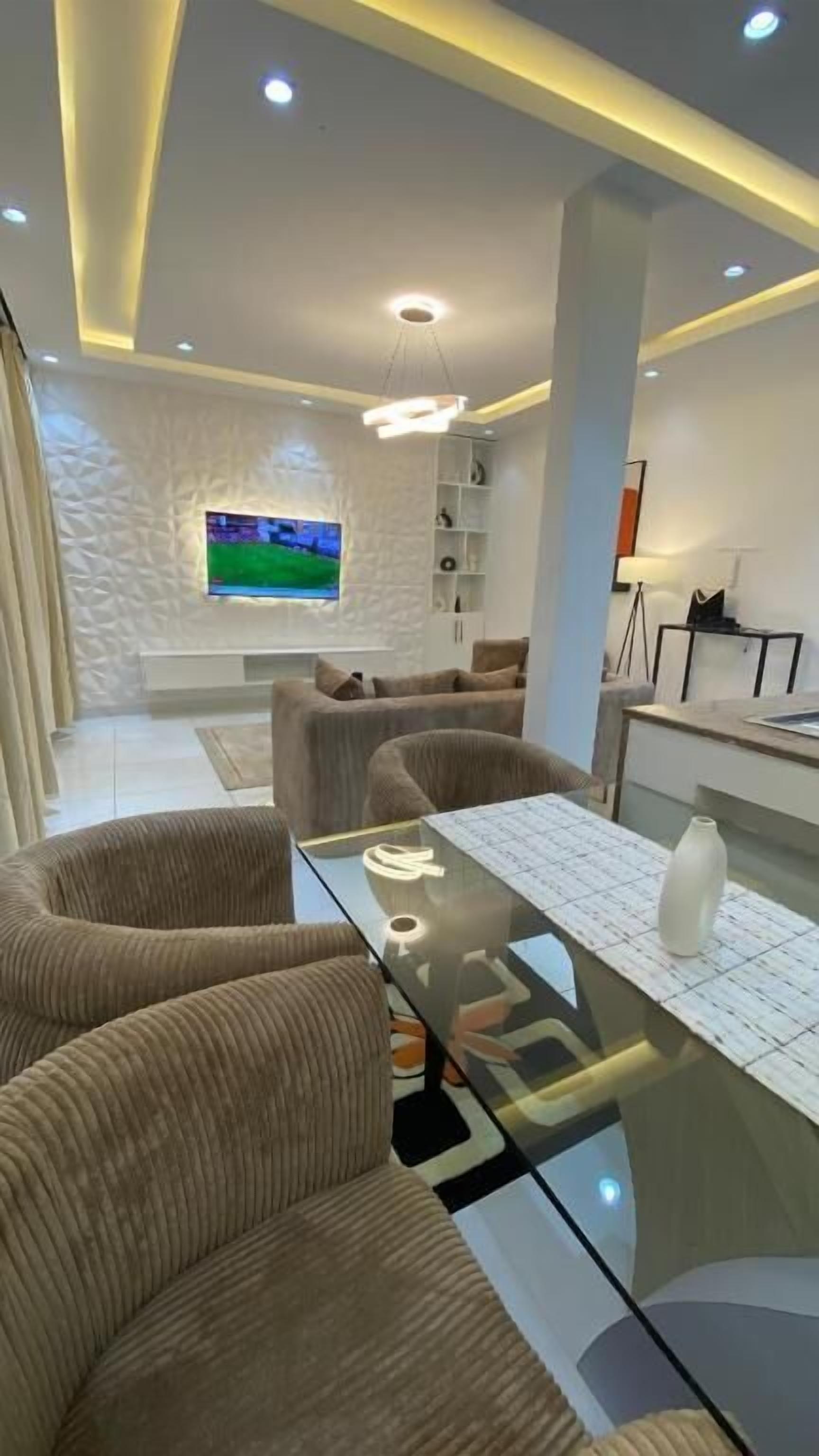 A modern living room with a glass coffee table, beige sofas, a TV mounted on the wall, and a ceiling with recessed lighting.