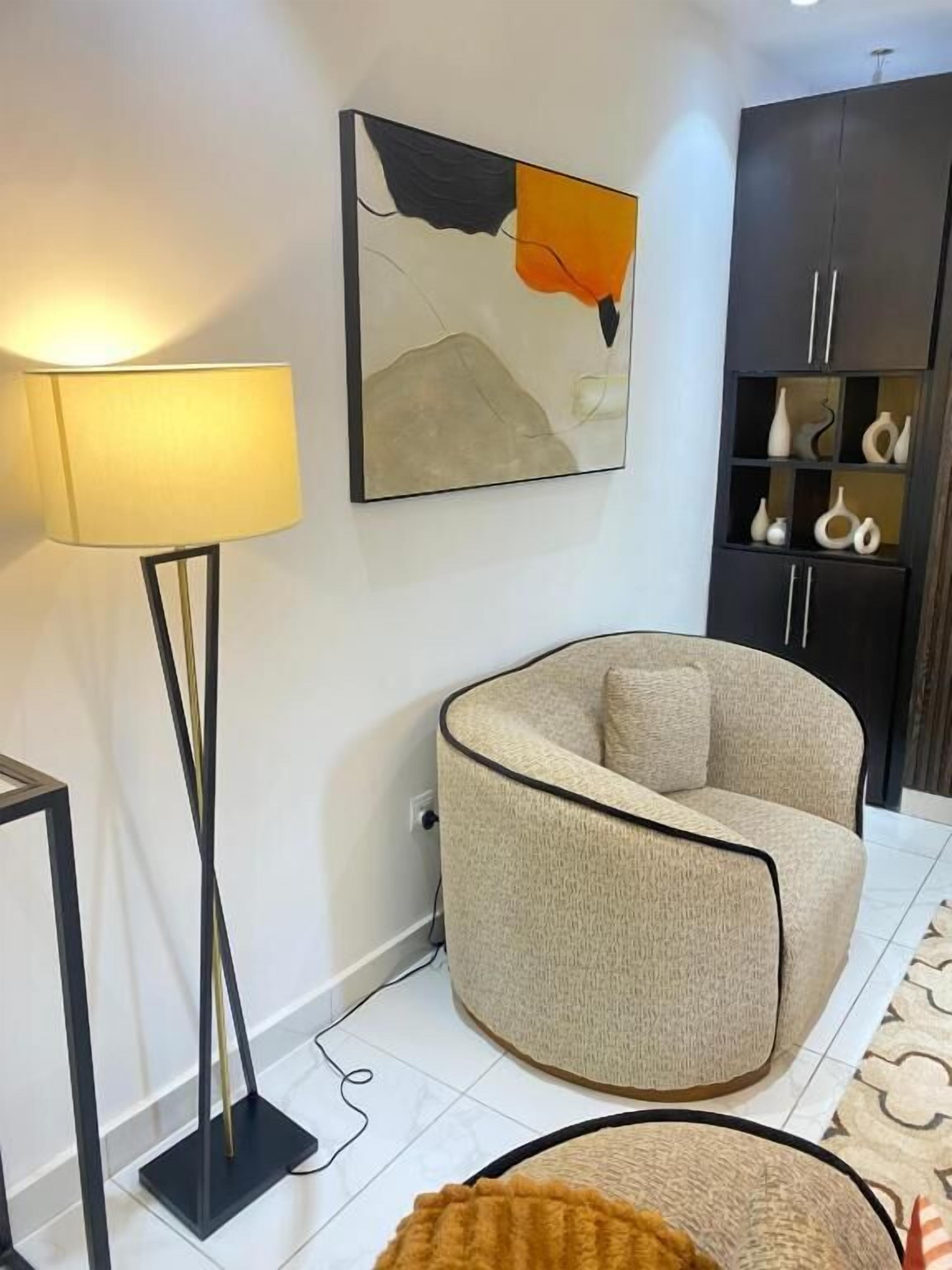 A room with a curved armchair, a floor lamp, a painting, and a cabinet with shelves.