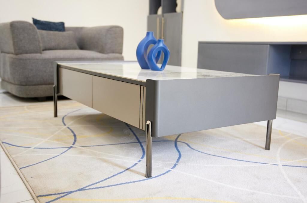 Una habitación moderna y minimalista con un sofá gris, una mesa de centro con superficie de vidrio y patas metálicas, y un jarrón azul.