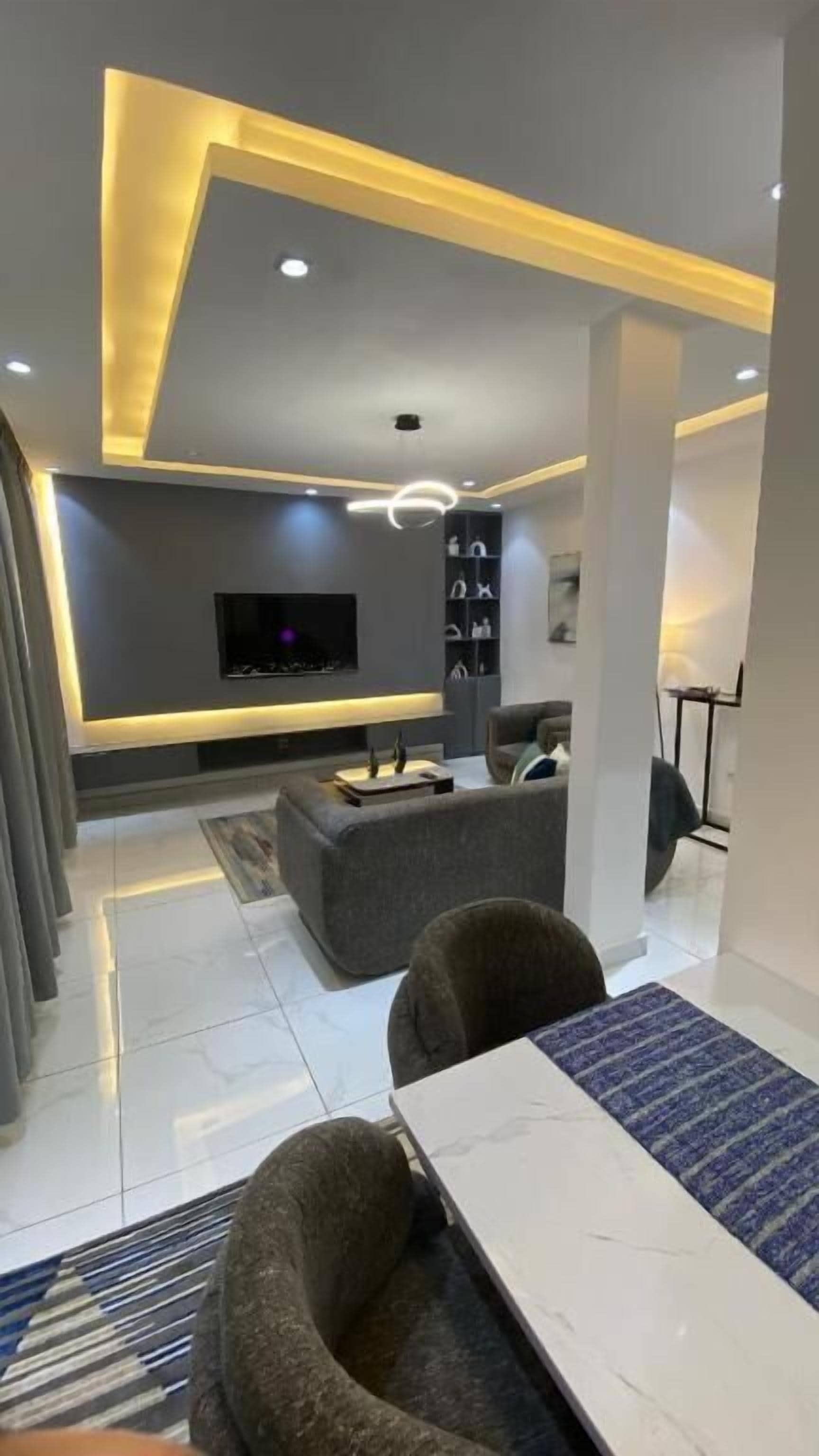 A modern living room with a flat-screen TV, grey sofas, a coffee table, and a dining area with a white table and chairs.