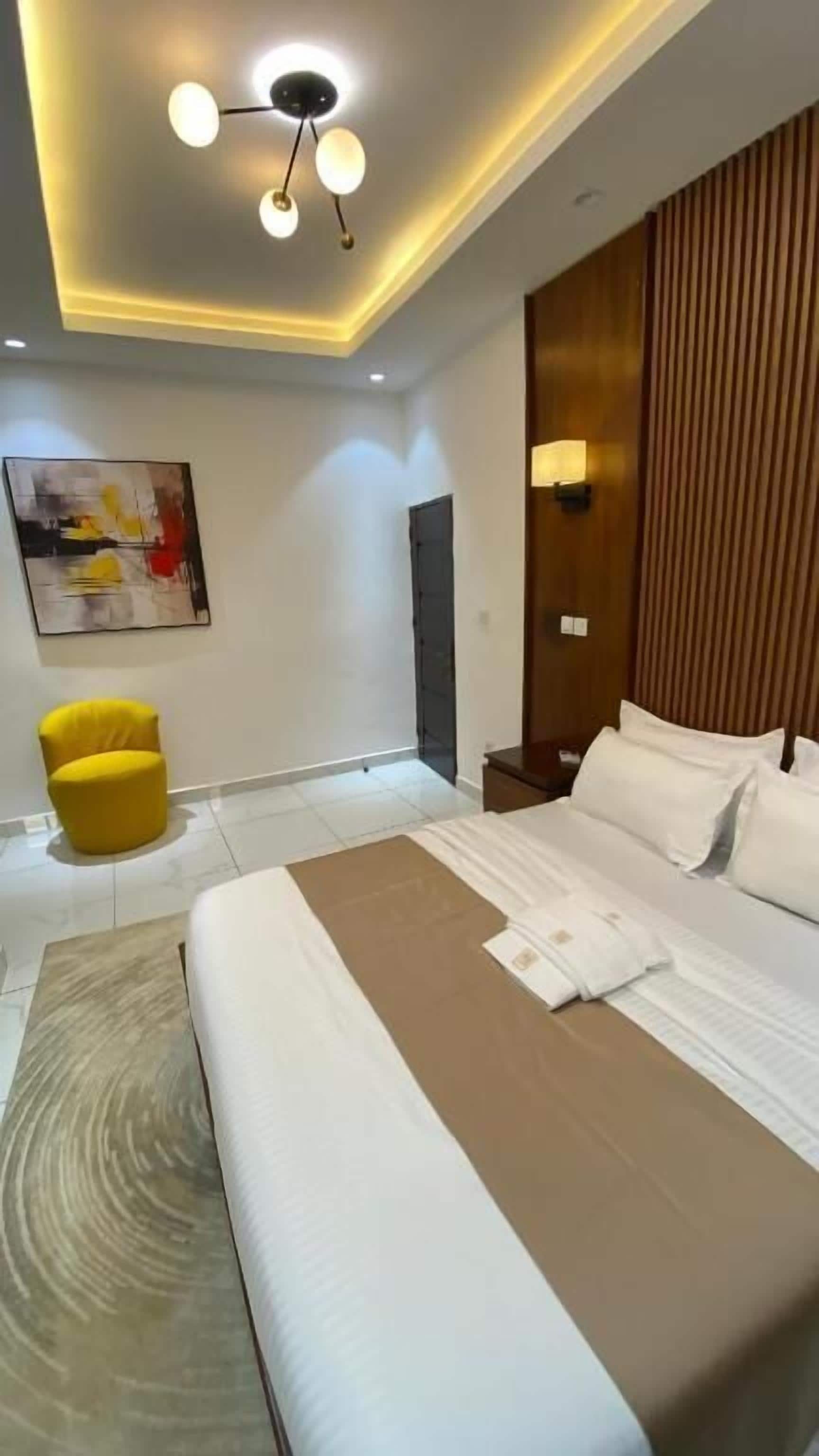 A modern hotel room with a large bed, a yellow chair, a painting on the wall, and a ceiling light fixture.
