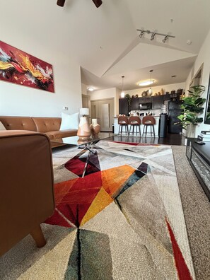 A living room with a sofa, a coffee table, and a colorful rug. There is a kitchen in the background with cabinets and a refrigerator. - Stylish 2BR/2BA OKC Apartment with Modern Decor, AC, Gym ,Pool & Plush Bedding (Oklahoma City)