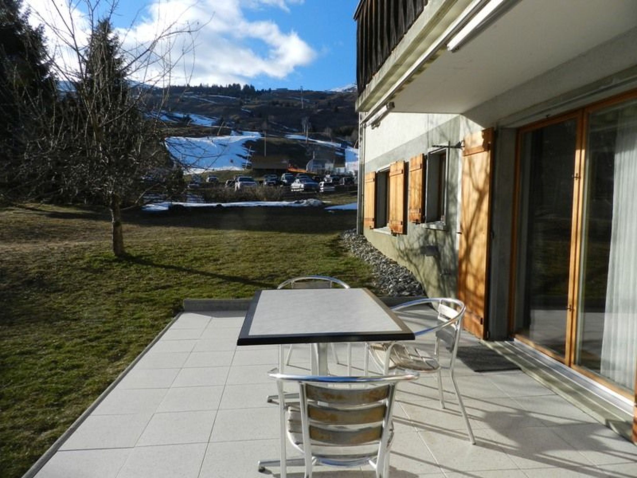 A patio with a table and chairs, a glass door leading to a building, and a view of a snowy landscape with trees and a mountain in the background.