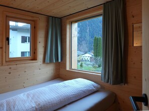 Una cama individual en una habitación tipo cabaña, con ventana que ofrece vistas a una montaña y un edificio.