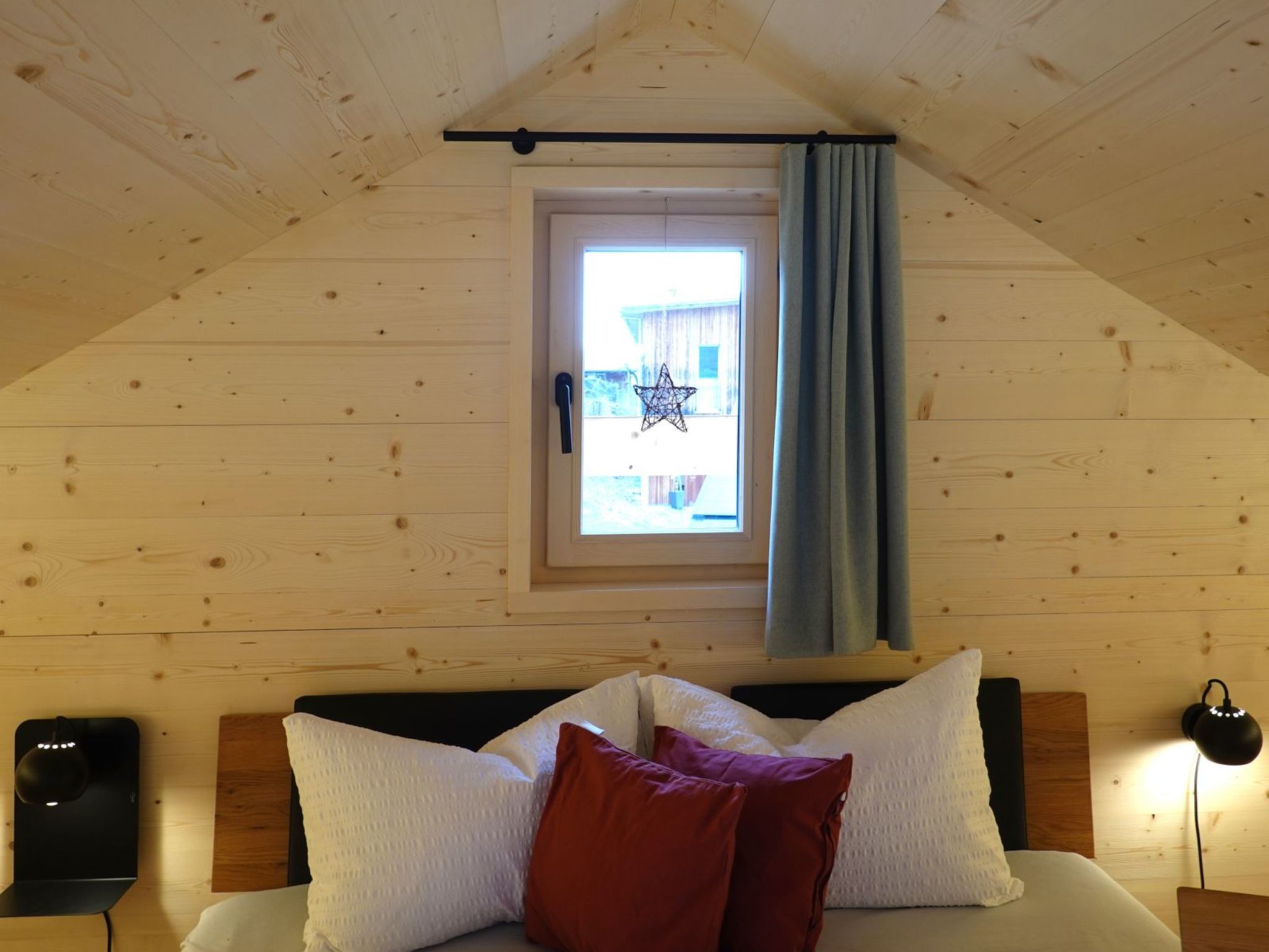 Ein Schlafzimmer mit schrägen Wänden, einem Fenster mit Vorhang, einem Bett mit weißen und roten Kissen sowie einem Nachttisch mit Lampe.