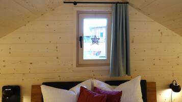 Ein Schlafzimmer mit schrägen Wänden, einem Fenster mit Vorhang, einem Bett mit weißen und roten Kissen sowie einem Nachttisch mit Lampe.