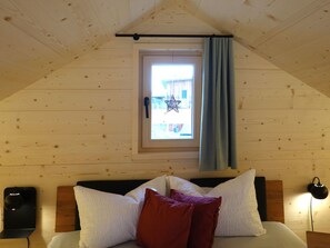 A bedroom with a sloped ceiling, a window with a curtain, a bed with white and red pillows, and a bedside table with a lamp.