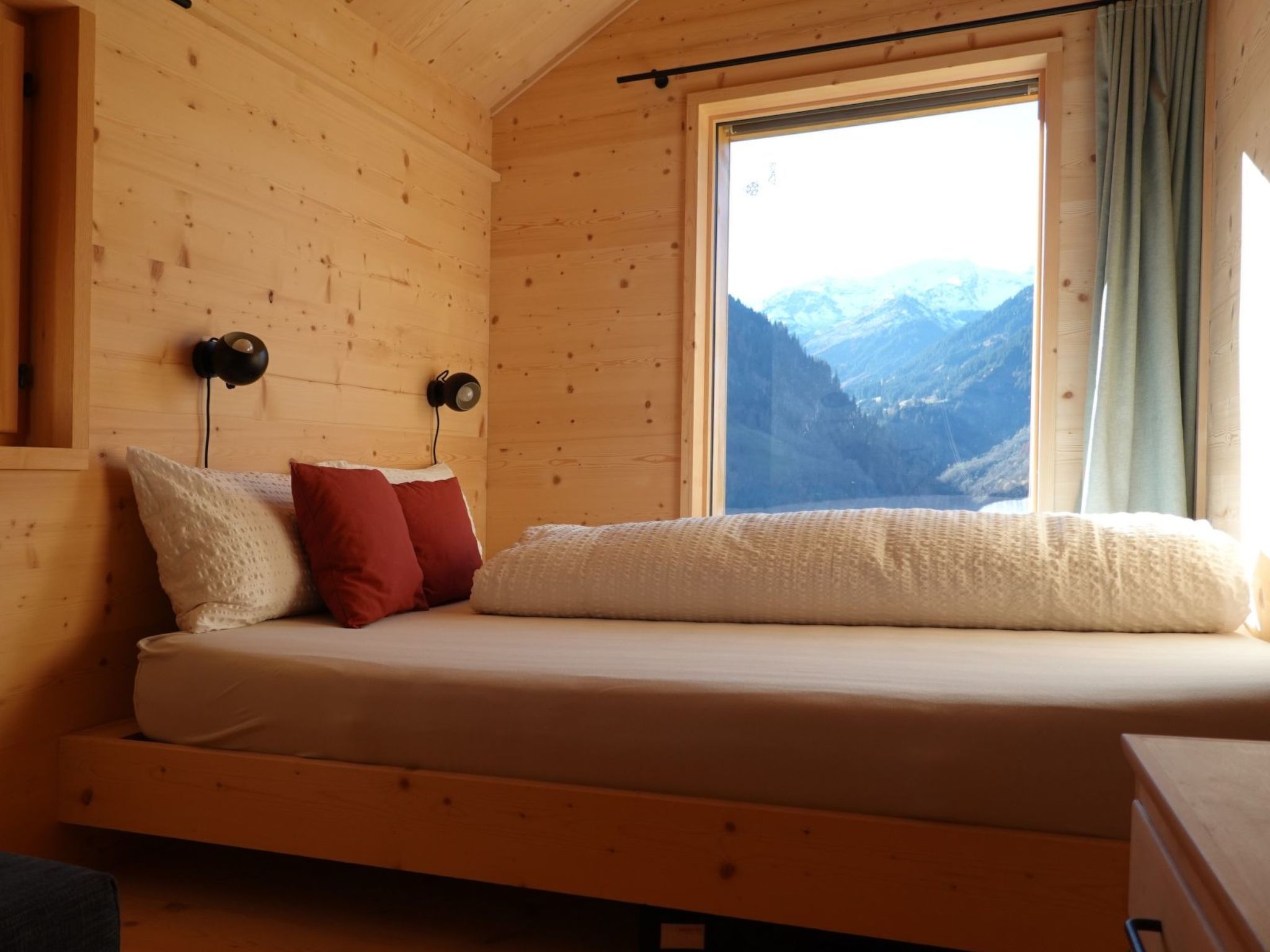 Ein Zimmer einer Holzhütte mit einem Bett, zwei Wandlampen und einem Fenster mit Blick auf die Berge.