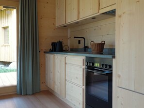 A kitchen with wooden cabinets, a black stove, and a window with a curtain. - Ferienhäuser Stiva Pintga, Rabius, (Rabius), 72010B by Interhome (Cumpadials)