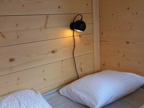A wooden cabin interior with a bed, pillows, and a wall-mounted light fixture. - Ferienhäuser Stiva Pintga, Rabius, (Rabius), 72010B by Interhome (Cumpadials)