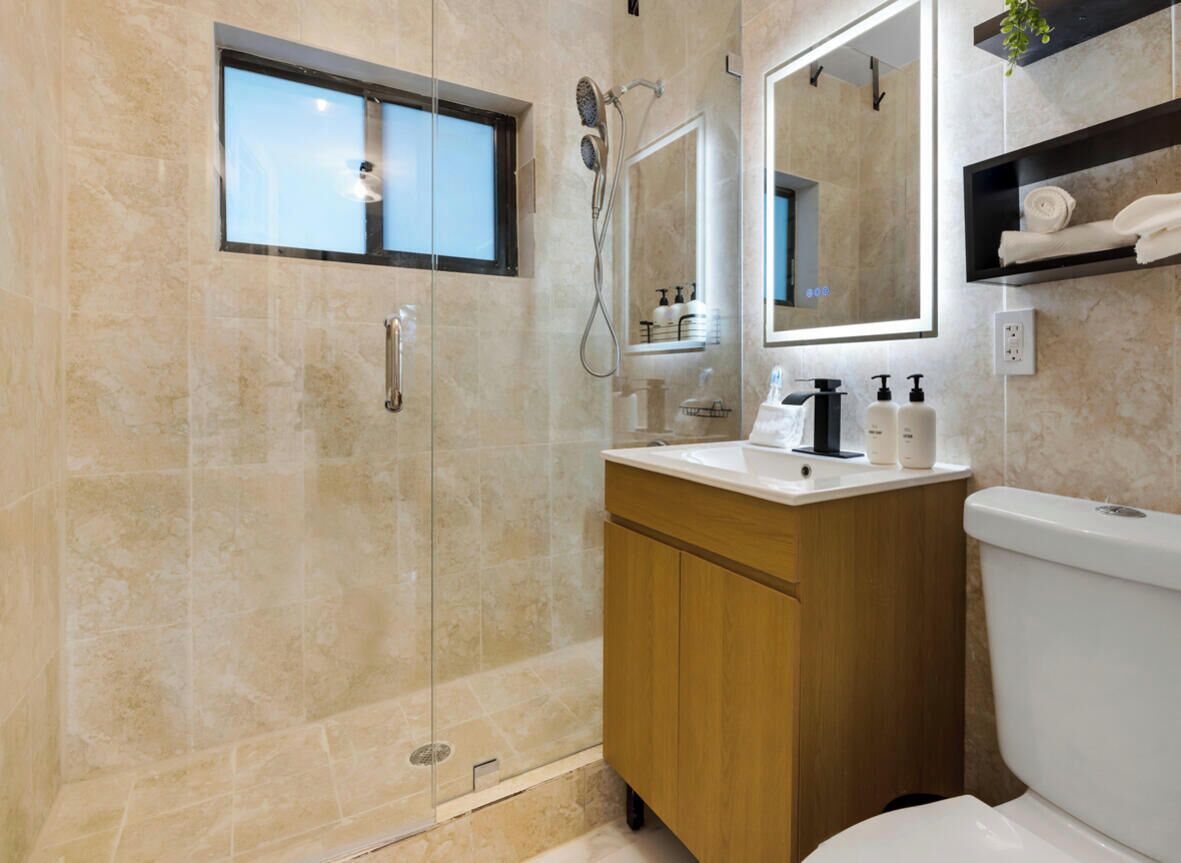Ein modernes Badezimmer mit einer gläsernen Duschkabine, einem Holzbadezimmer-Möbel, einer Toilette und einem Spiegel mit Ablagefläche darüber.