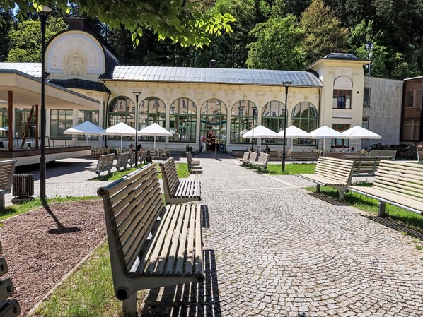 Ein gepflasterter Weg mit Bänken, Sitzgelegenheiten im Freien unter Sonnenschirmen und ein Gebäude mit Rundbogenfenstern im Hintergrund.