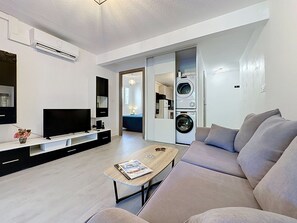A modern living room with a flat-screen TV, a sofa, a coffee table with magazines, a washing machine, and a kitchen area visible through an open door. - Dixie Land by Interhome (La Grande Motte)