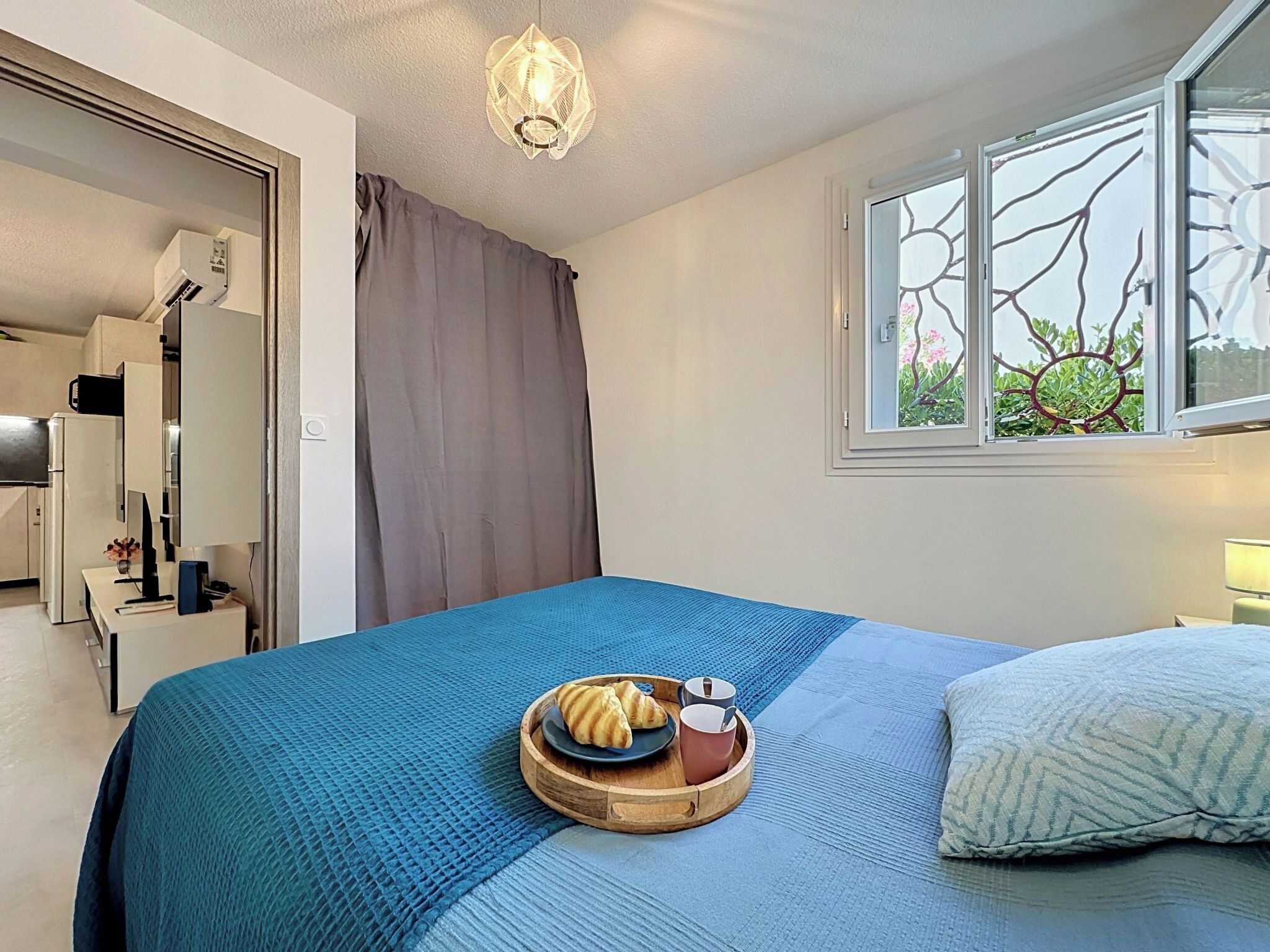 Un dormitorio con cama, una mesita de noche con una bandeja de croissants y tazas de café, una ventana con vistas a árboles y una lámpara en el techo.
