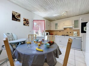 A dining area with a table set for breakfast, a chair, and a kitchen in the background. - L'Estuaire by Interhome (Saint Brevin les Pins)