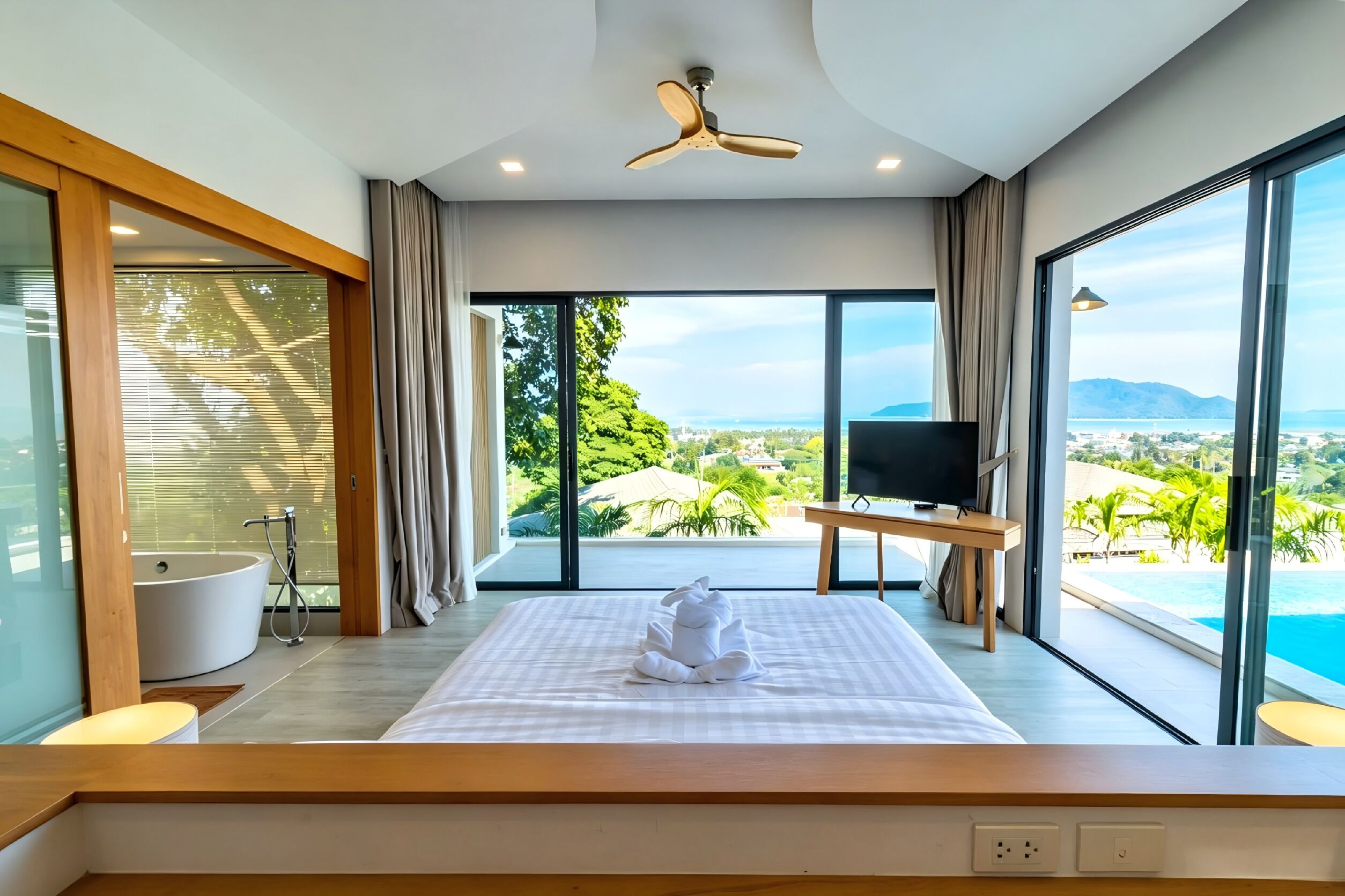 A modern hotel room with a large bed, a ceiling fan, a TV, and a view of the ocean.