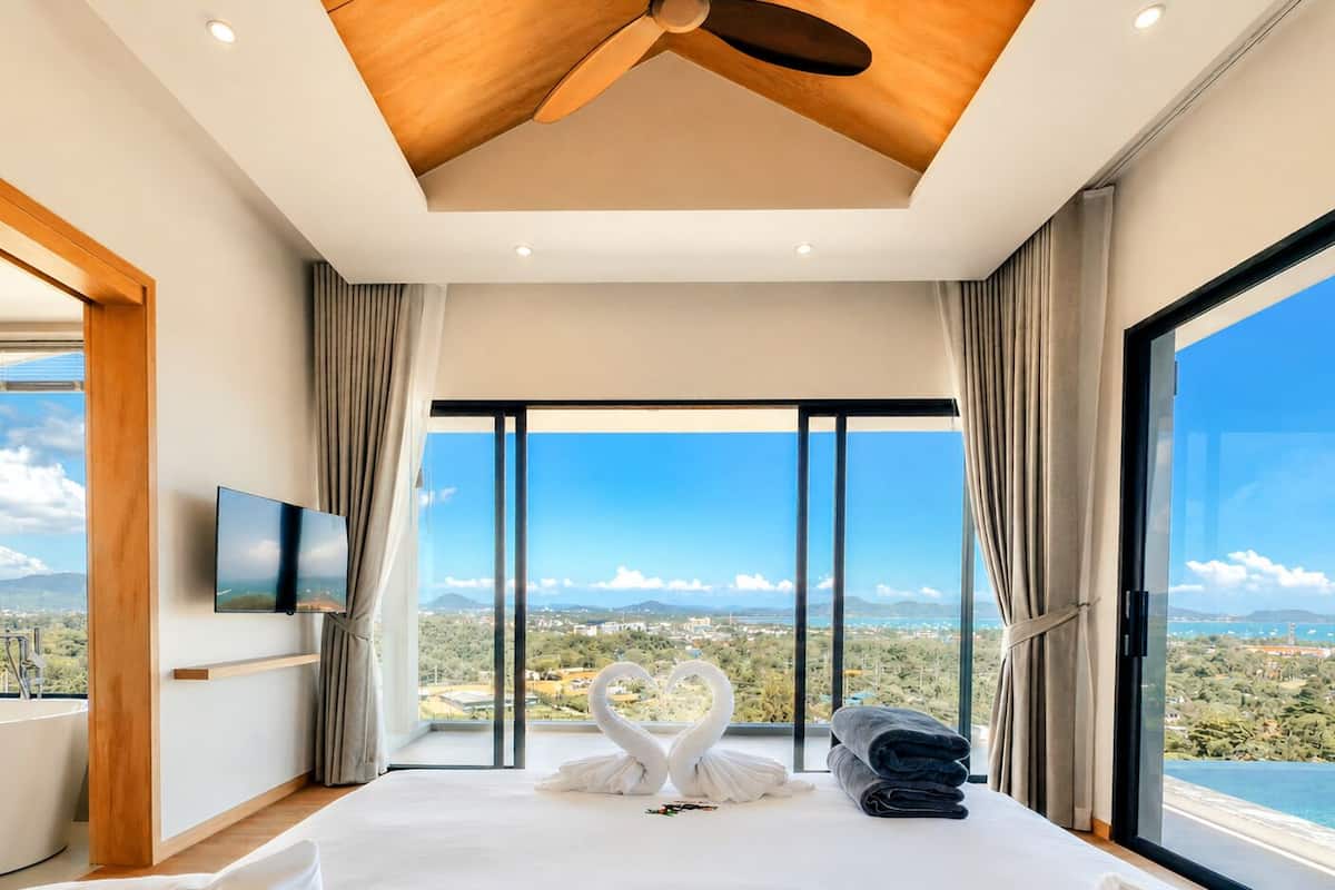 Ein modernes Schlafzimmer mit einem großen Bett, einem Deckenventilator, einem Flachbildfernseher und Blick auf eine Küstenlandschaft.