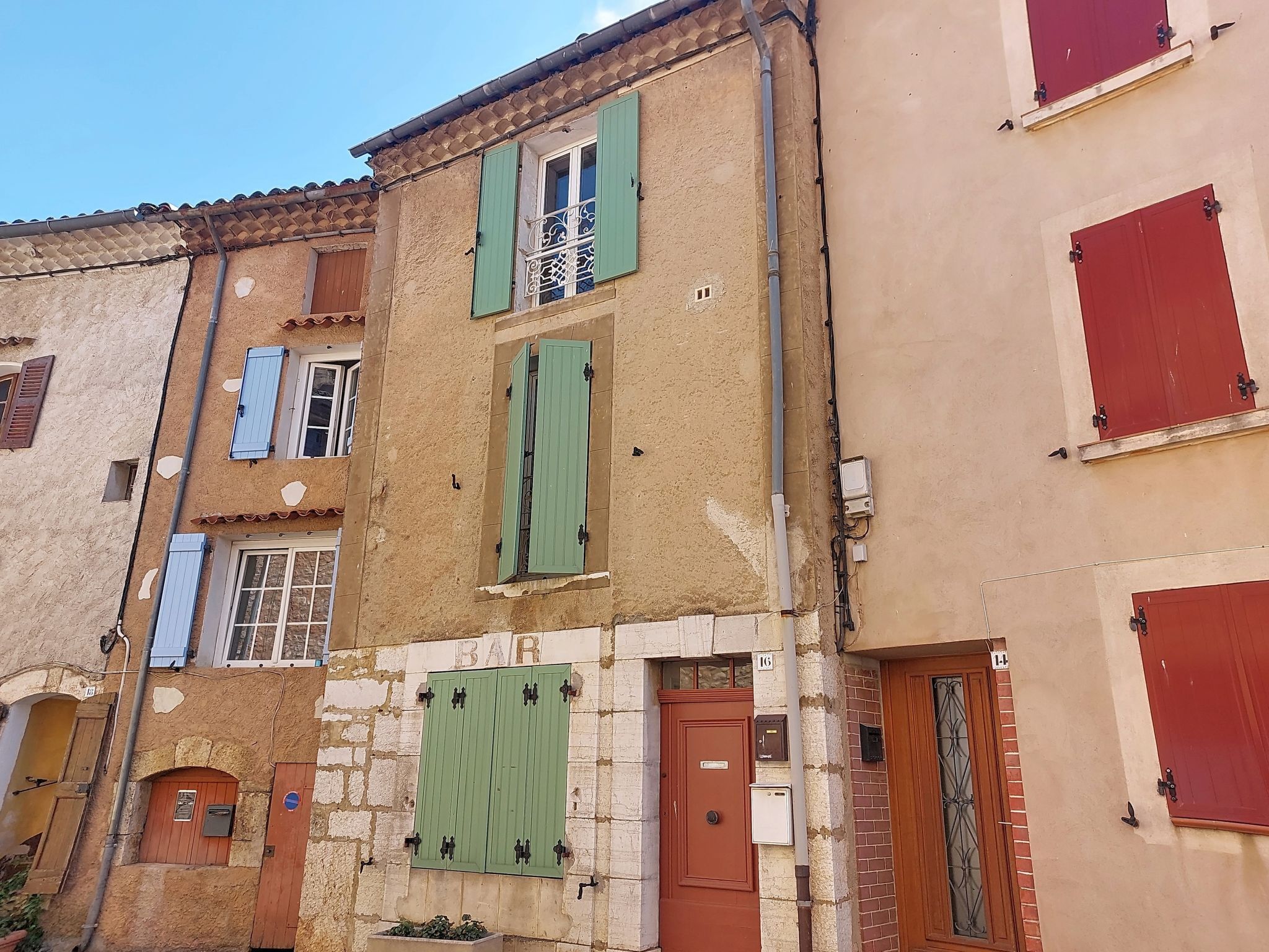 Un immeuble à plusieurs étages, dont la façade est un mélange de bois et de pierre, avec des volets verts et une porte bien visible portant l’inscription « BAR ».
