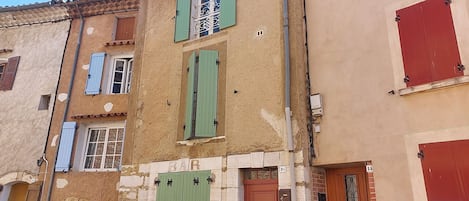 Un immeuble à plusieurs étages, dont la façade est un mélange de bois et de pierre, avec des volets verts et une porte bien visible portant l’inscription « BAR ».
