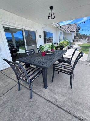 Eine Terrasse im Freien mit Tisch und Stühlen, umgeben von Blumentöpfen und einem weißen Gebäude mit großen Fenstern.