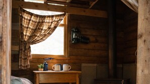 A cozy cabin interior with a wooden bed, a small table with mugs, a window with plaid curtains, a lantern, and a wood-burning stove. - Spruce, the tiny cabin in the woods (Markdale)
