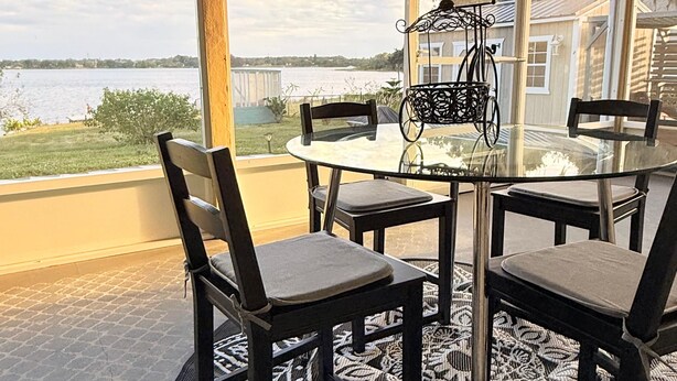 A round table with chairs and a patterned rug on a balcony with a view of a lake and buildings.