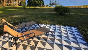 A patio with a checkered floor and lounge chairs, a grassy area with trees, and a body of water in the background.